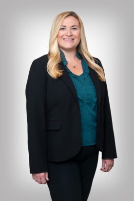 A woman in black jacket and blue shirt standing next to wall.